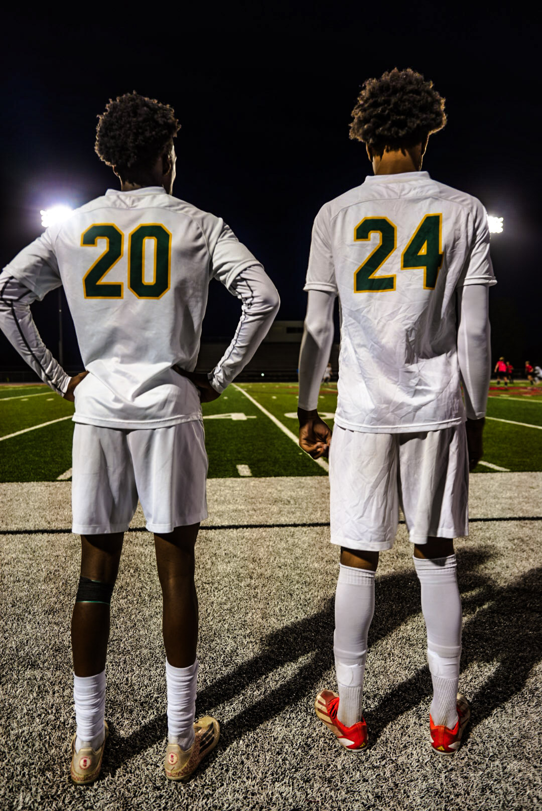 Two soccer players numbered 20 and 24 standing side by side on the sideline at night