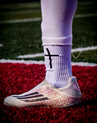 Close up of a soccer sock with a hand-drawn cross and cleats on red turf
